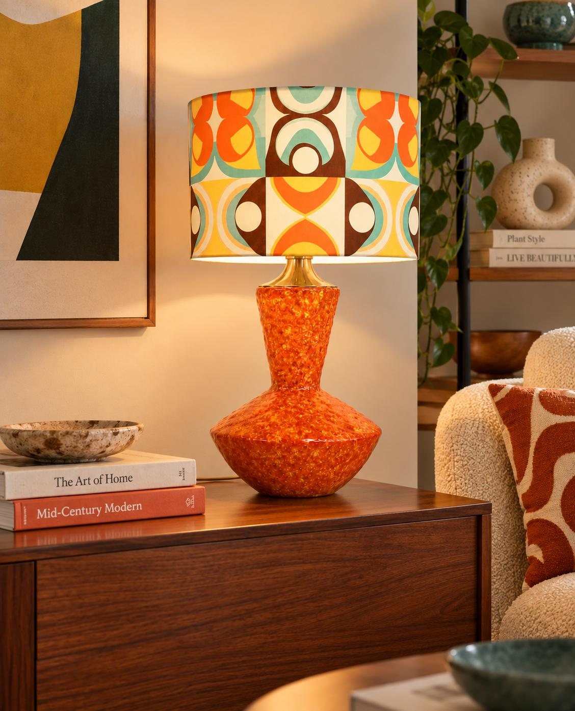 A Lamparita lamp with a retro geometric-print shade, lit on a mid-century sideboard beside books, a plant, and framed art